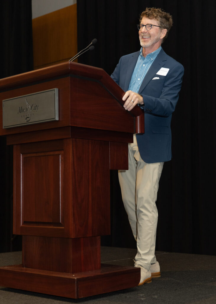 An individual wearing glasses and a blue blazer stands smiling at a podium while speaking at an event. The podium has a plaque labeled 'Music City Center,' and the background features a dark curtain.