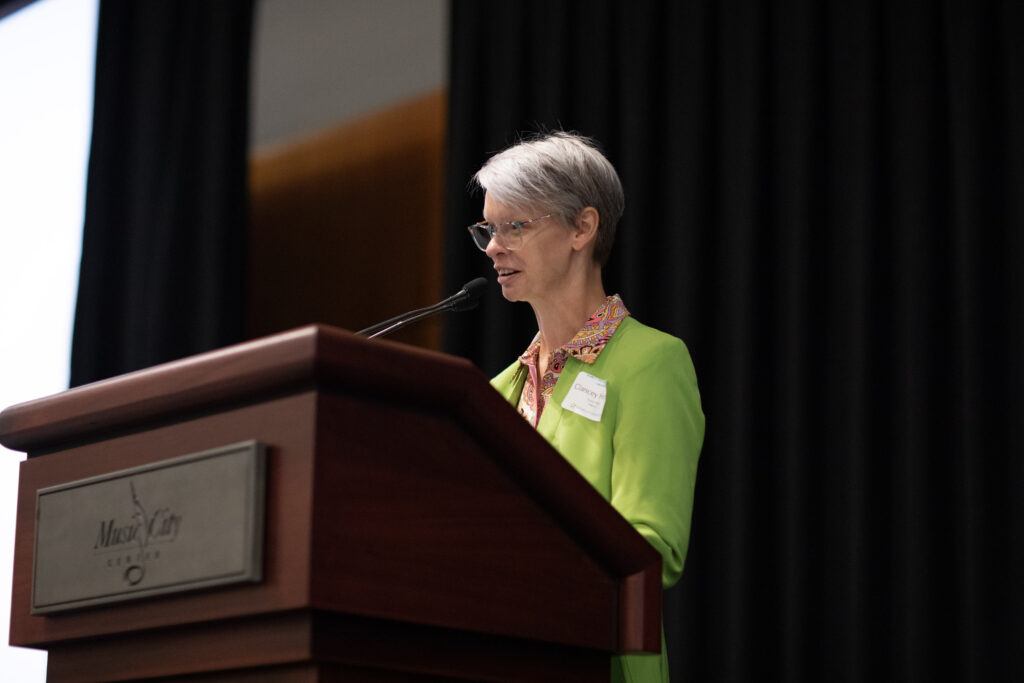 The image shows a person with short gray hair speaking at a podium. They are wearing glasses and a bright green jacket with a colorful collar. The background features a dark, heavy curtain, indicating a conference hall. The wooden podium has a gray plaque on the front.