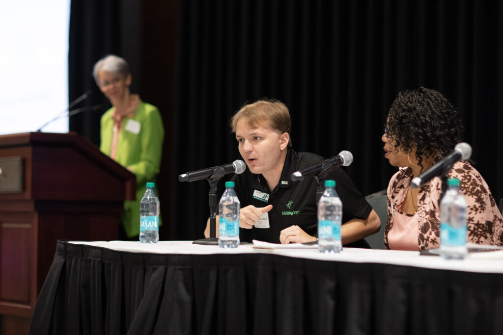 A man speaks into a microphone with a woman next to him, engaged in discussion. Several water bottles are visible on the table. A person stands at a podium in the background, slightly blurred. The setting is a conference.